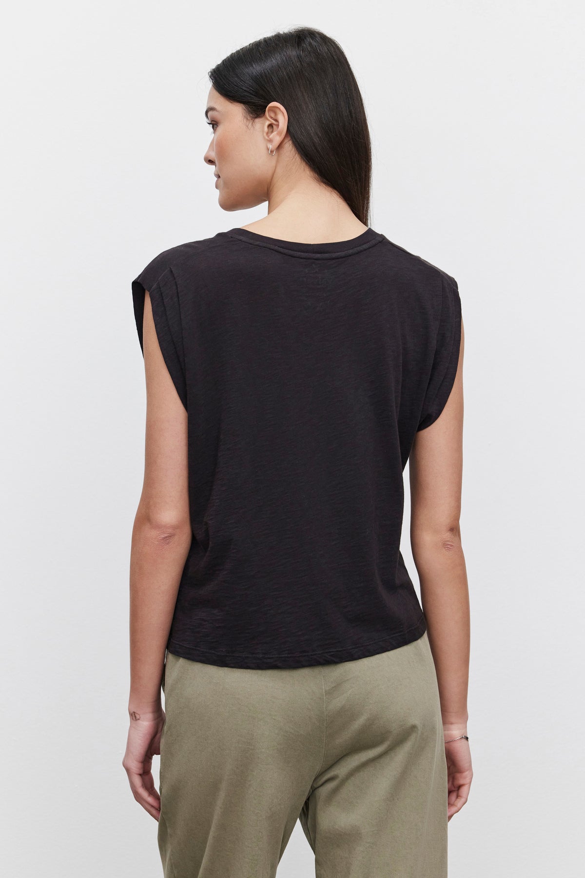   A person with long hair wearing a sleeveless black Cecile Tank Top from Velvet by Graham & Spencer, made from soft cotton slub and olive green pants is standing, facing away from the camera. The tank top features a subtle pleated shoulder detail, adding an elegant touch to the casual ensemble. 