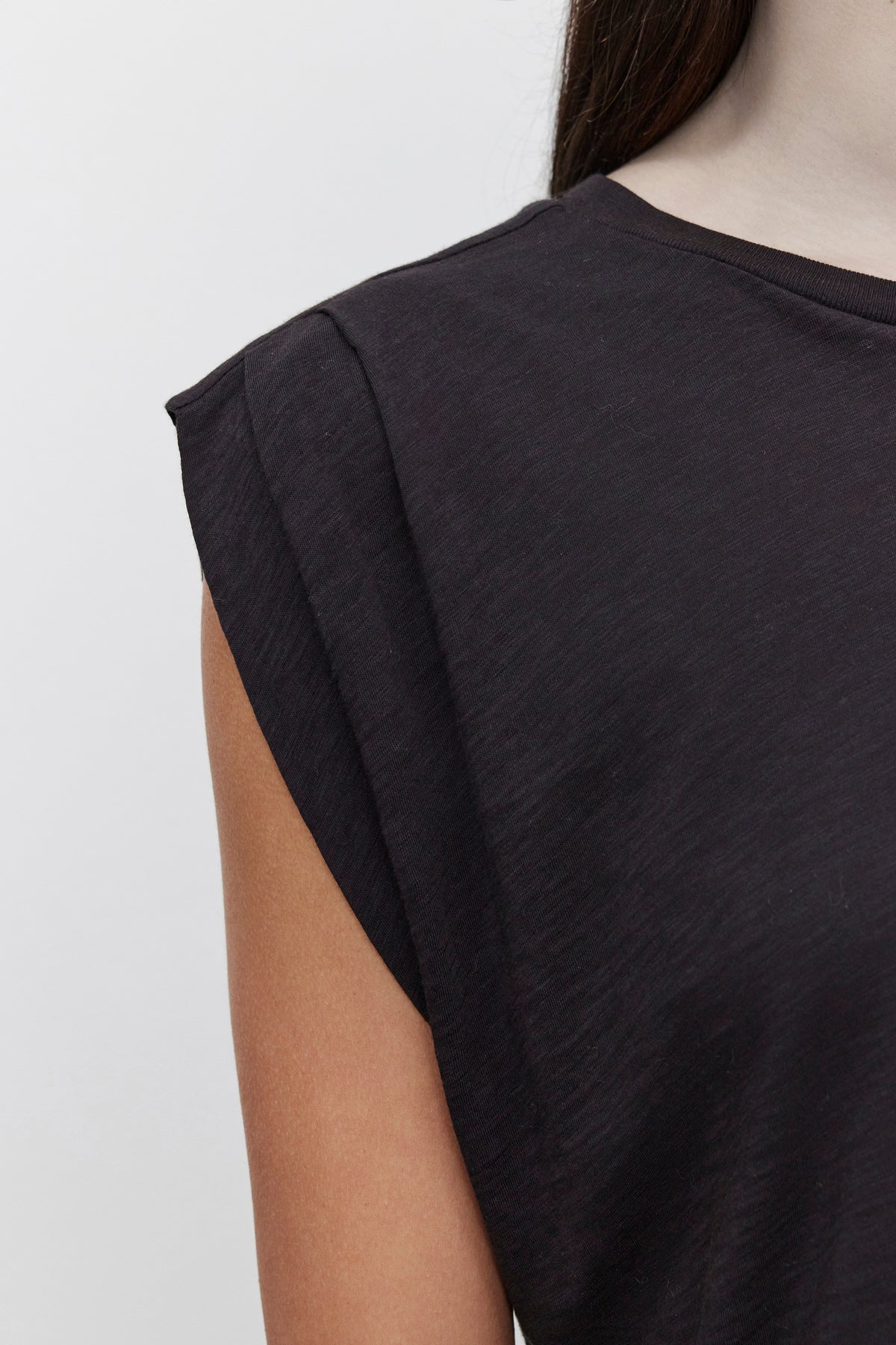   Close-up of a person wearing the CECILE TANK TOP by Velvet by Graham & Spencer in black. The sleeveless shirt, made from soft cotton slub fabric, showcases the person's arm and shoulder against a plain background, featuring subtle pleated shoulder details for added flair. 