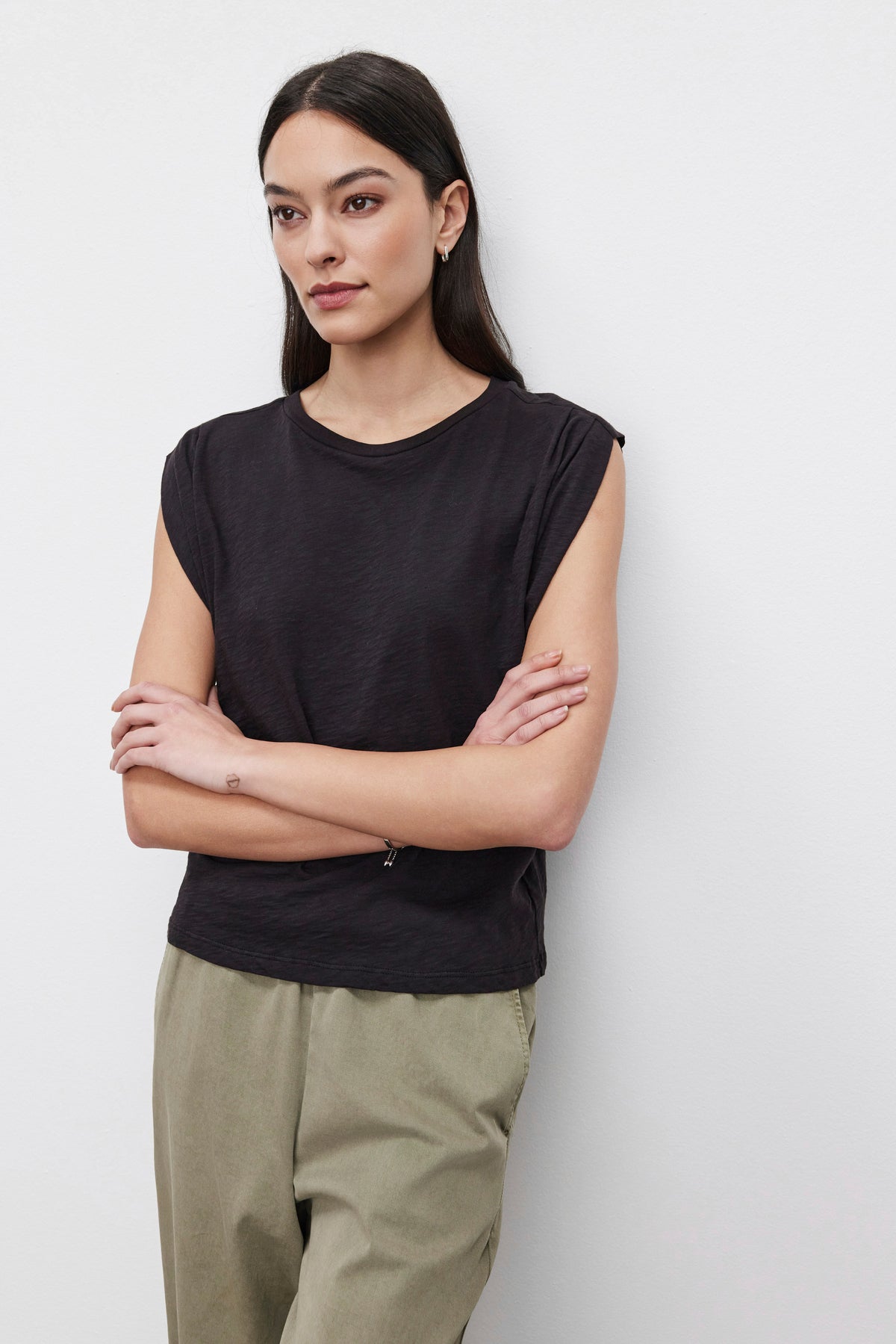   A person with long hair stands against a plain background, wearing a sleeveless black CECILE TANK TOP by Velvet by Graham & Spencer, featuring pleated shoulder detail and beige pants, arms crossed. 