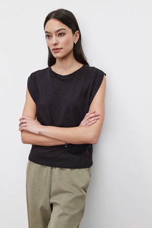 A person with long hair stands against a plain background, wearing a sleeveless black CECILE TANK TOP by Velvet by Graham & Spencer, featuring pleated shoulder detail and beige pants, arms crossed.