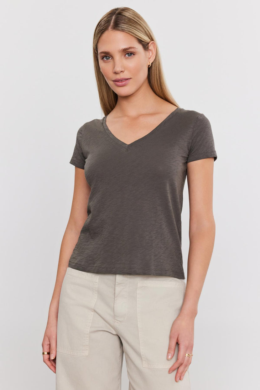 A woman in a Velvet by Graham & Spencer ELIAH TEE and beige pants standing against a white background.