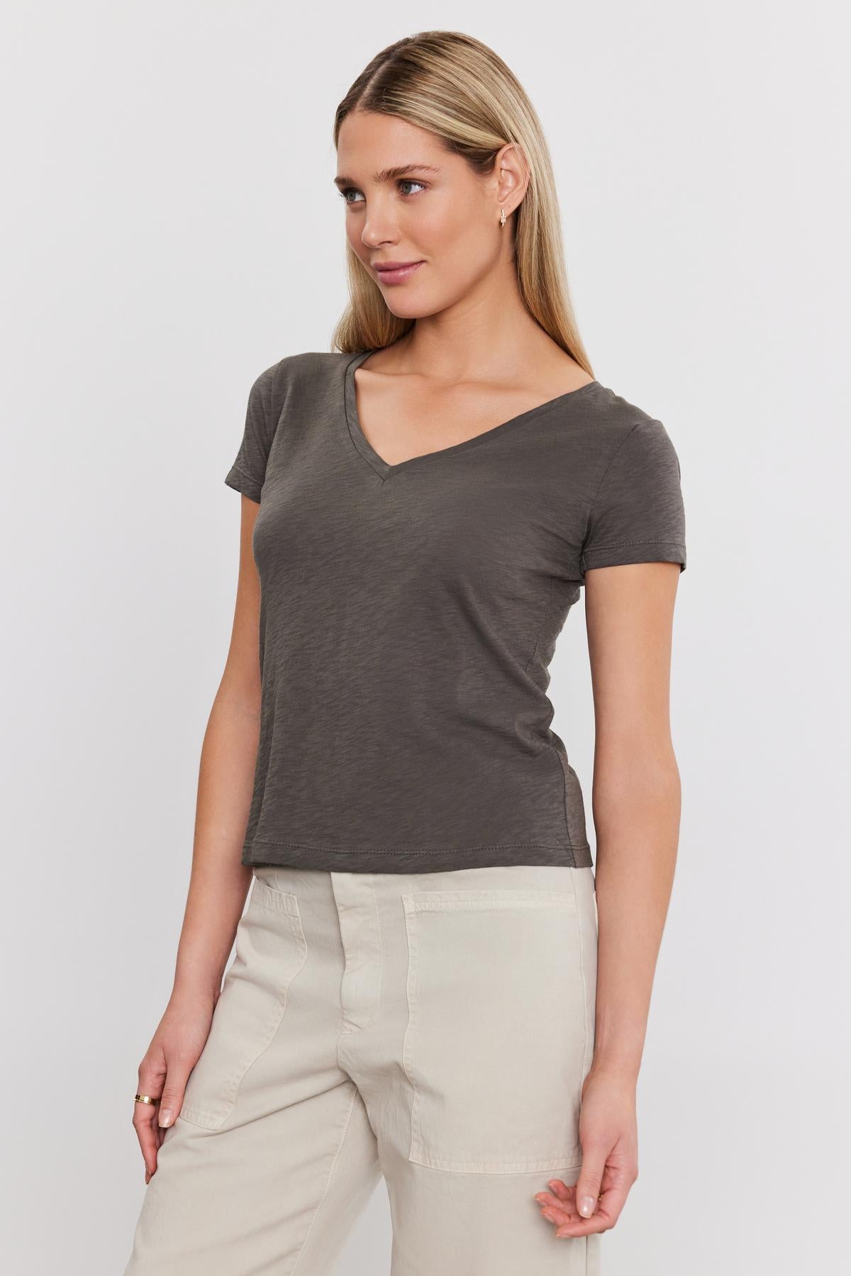 A woman in a gray Eliah Tee from Velvet by Graham & Spencer with a v-neckline and beige trousers, standing against a white background, looking slightly to her right.-36910213529793