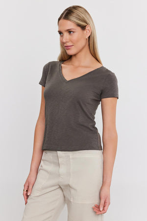 A woman in a gray Eliah Tee from Velvet by Graham & Spencer with a v-neckline and beige trousers, standing against a white background, looking slightly to her right.