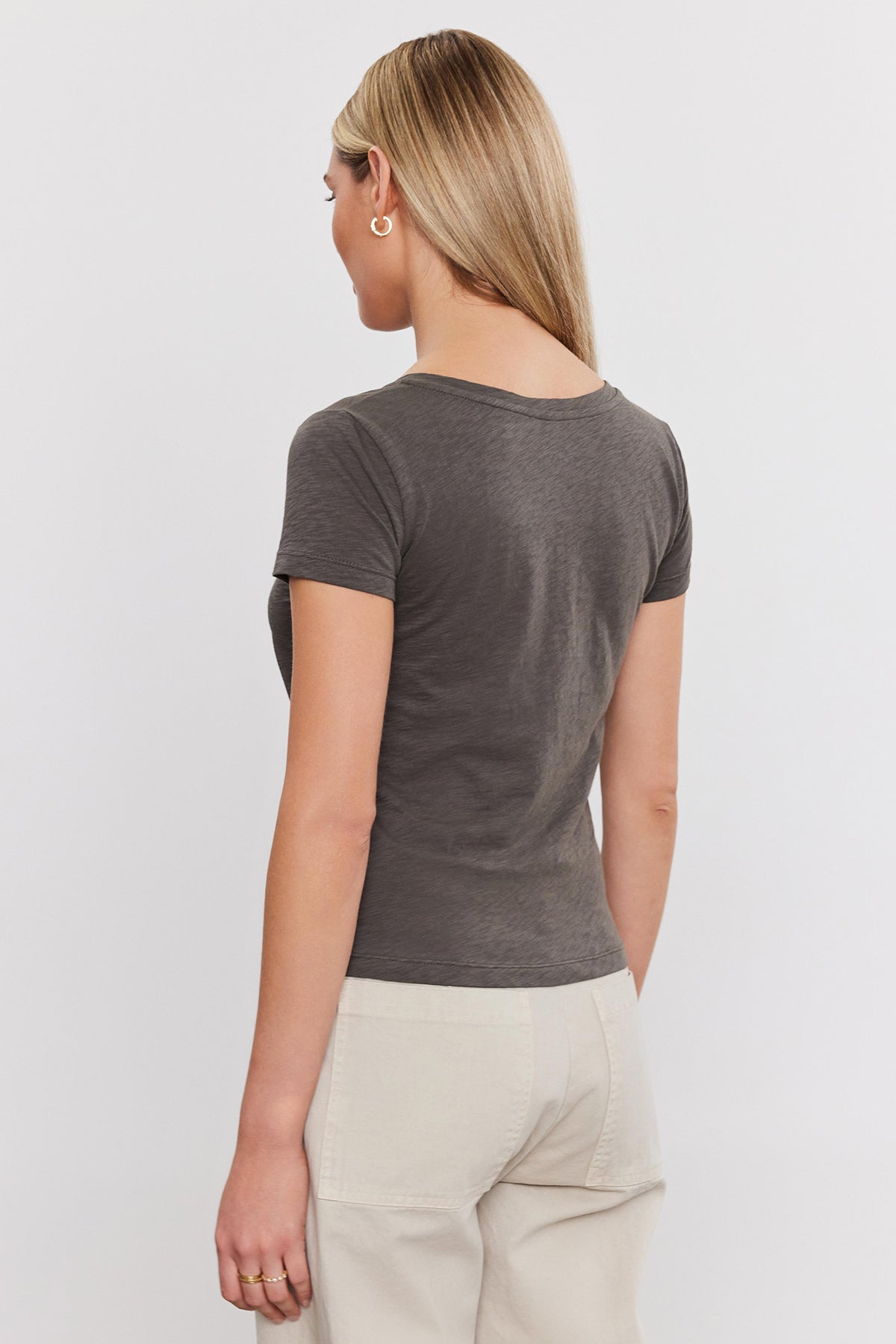   A woman facing away, wearing a Velvet by Graham & Spencer ELIAH TEE t-shirt and beige pants, standing against a plain white background. 