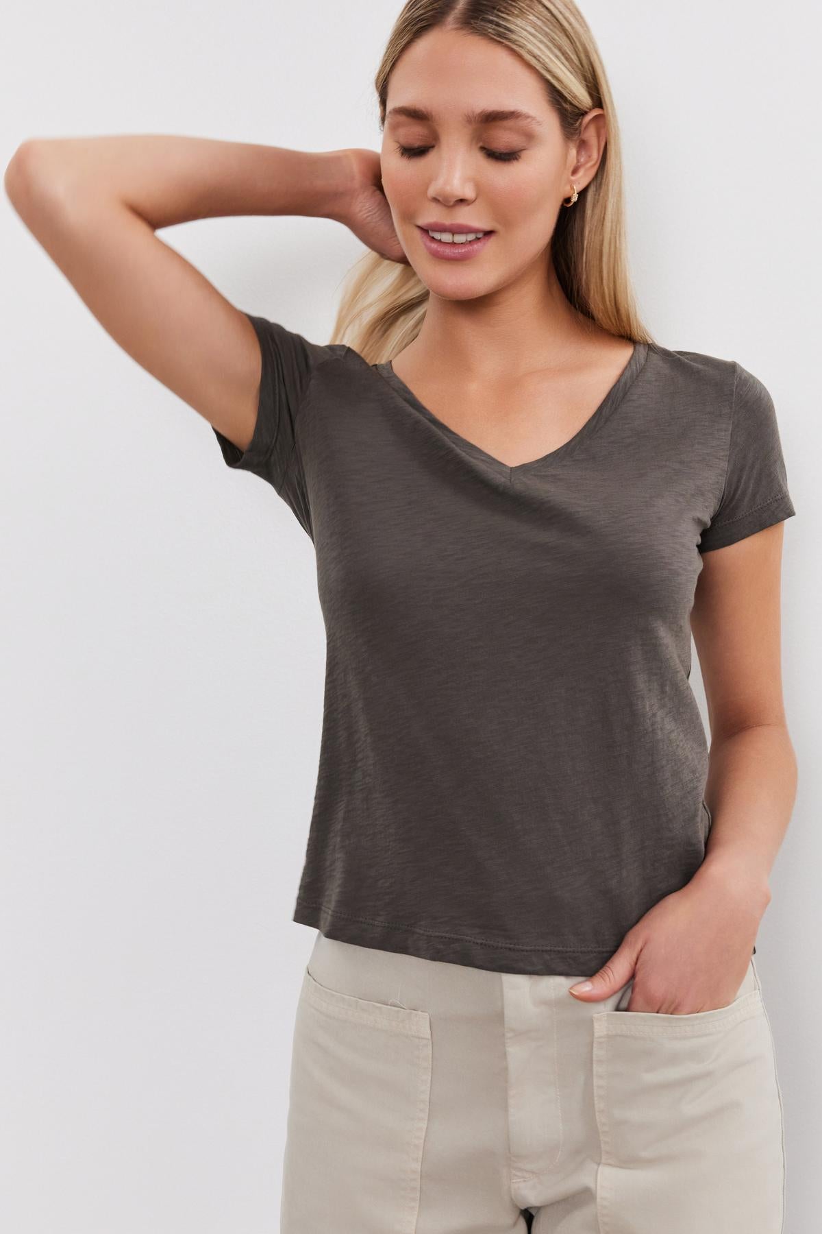   Woman in a gray Velvet by Graham & Spencer ELIAH TEE and beige pants, posing with her hand on her head, smiling gently. 