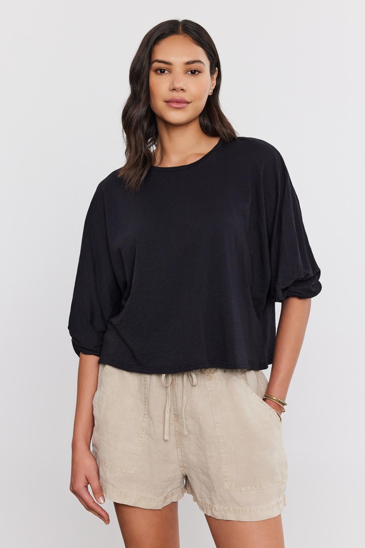   A woman stands wearing a black cotton slub FAITH TEE and beige drawstring shorts, posing against a white background by Velvet by Graham & Spencer. 