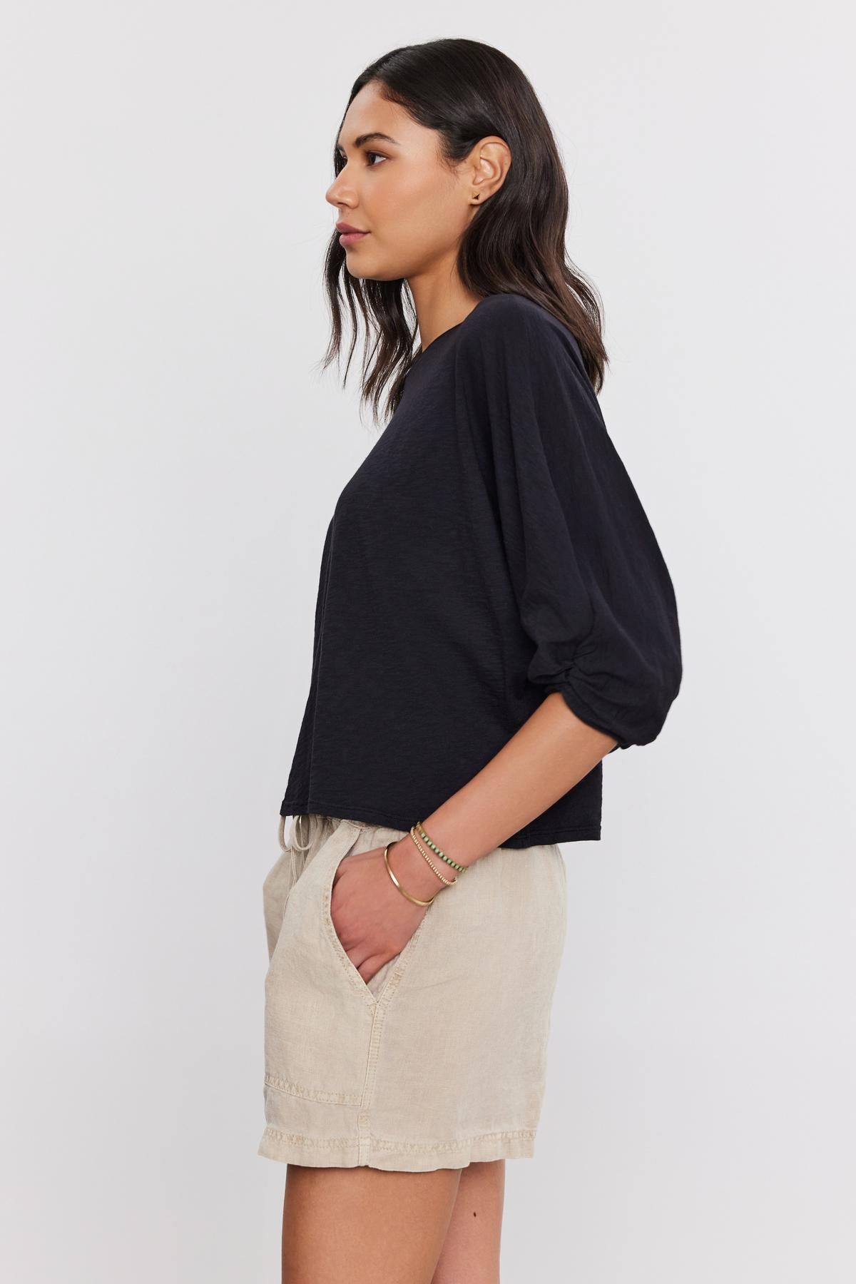   Profile view of a woman with shoulder-length dark hair, wearing a black Velvet by Graham & Spencer FAITH TEE and beige shorts, standing against a white background. 