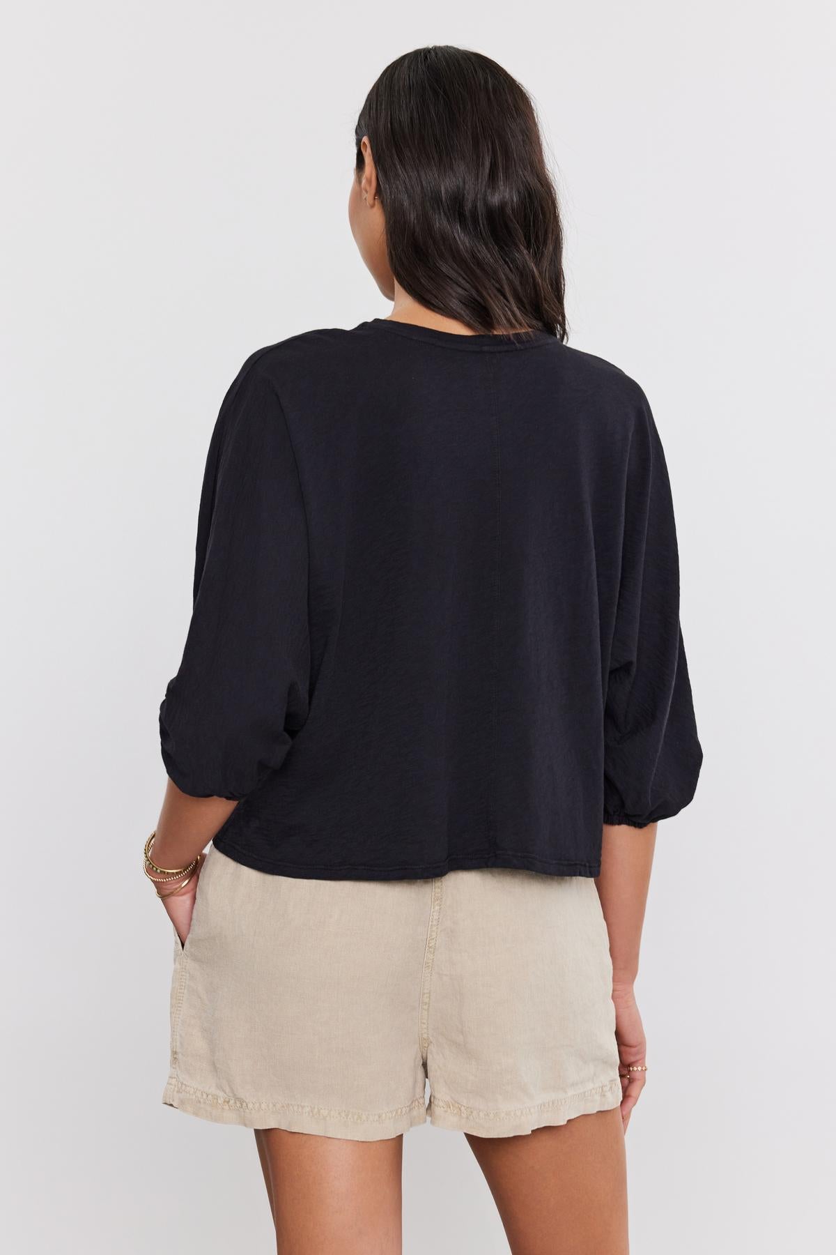   Rear view of a woman wearing a black FAITH TEE by Velvet by Graham & Spencer with dolman sleeves and beige, high-waisted shorts, facing away from the camera. 