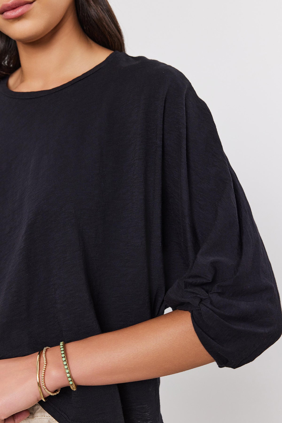   Close-up of a woman in a black blouse with dolman sleeves, focusing on the upper body and arms, wearing gold bracelets. (Product: FAITH TEE by Velvet by Graham & Spencer) 