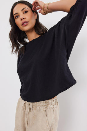 A woman in a black Velvet by Graham & Spencer FAITH TEE and beige drawstring pants, posing with one arm raised.