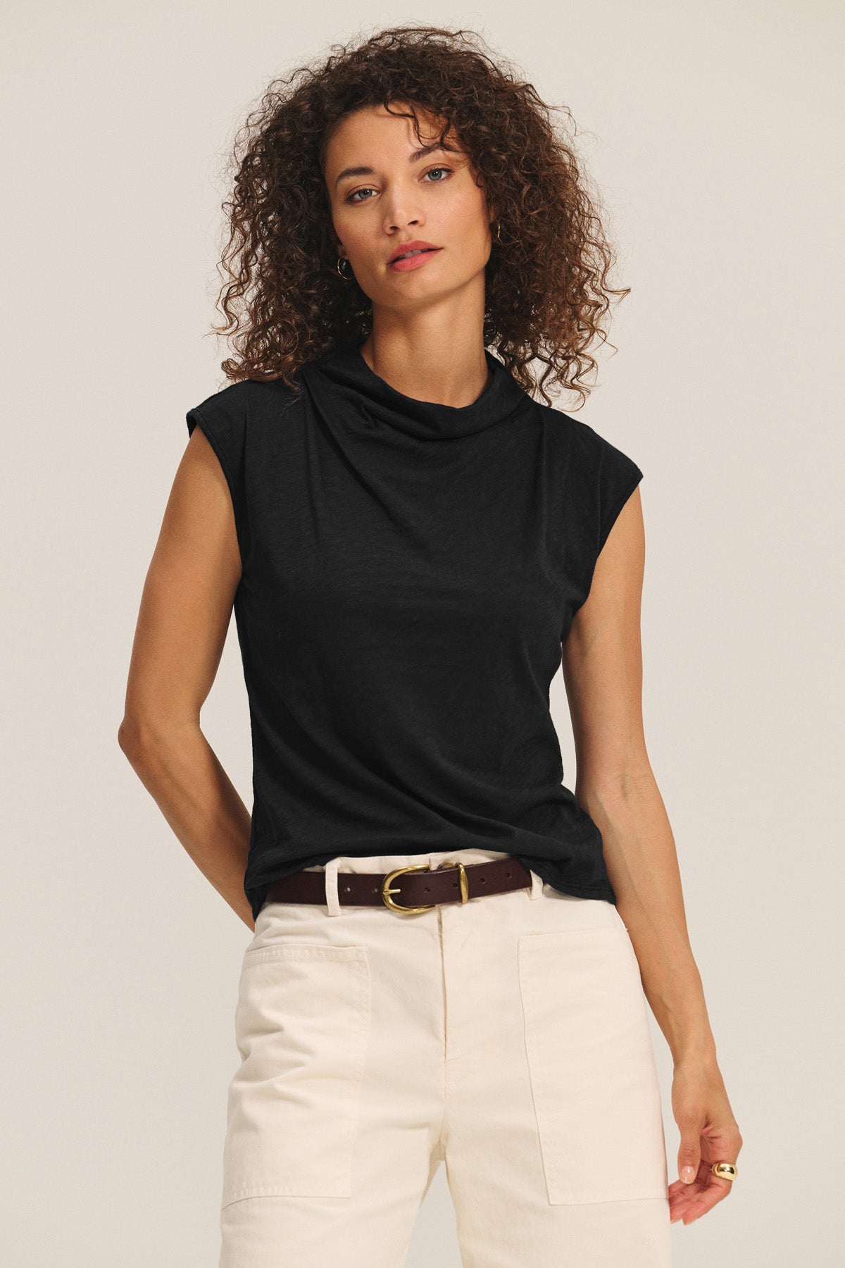 A woman with curly hair wears the Velvet by Graham & Spencer FLETCHER TEE, a black sleeveless mock neck top, tucked into high-waisted white pants with a brown belt, standing against a plain background.-40946686165185