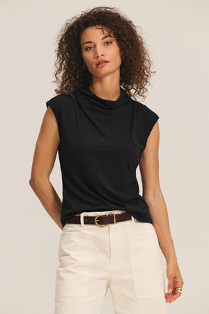 A woman with curly hair wears the Velvet by Graham & Spencer FLETCHER TEE, a black sleeveless mock neck top, tucked into high-waisted white pants with a brown belt, standing against a plain background.