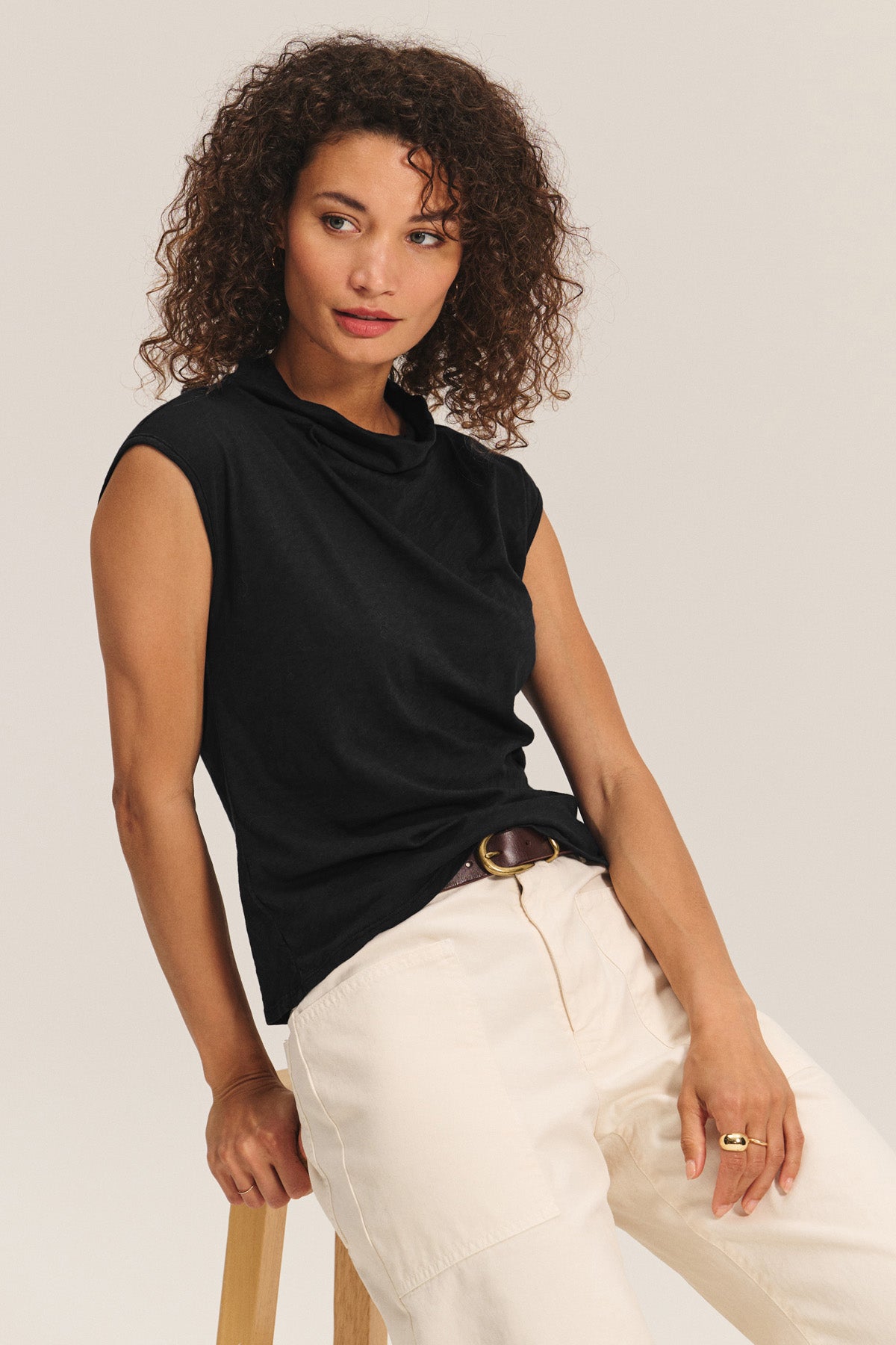 A woman with curly hair wears the FLETCHER TEE by Velvet by Graham & Spencer—a sleeveless mock neck top—paired with light pants, sitting on a wooden stool against a plain background. Ideal as a versatile layering piece for any look.