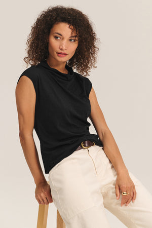 A woman with curly hair wears the FLETCHER TEE by Velvet by Graham & Spencer—a sleeveless mock neck top—paired with light pants, sitting on a wooden stool against a plain background. Ideal as a versatile layering piece for any look.