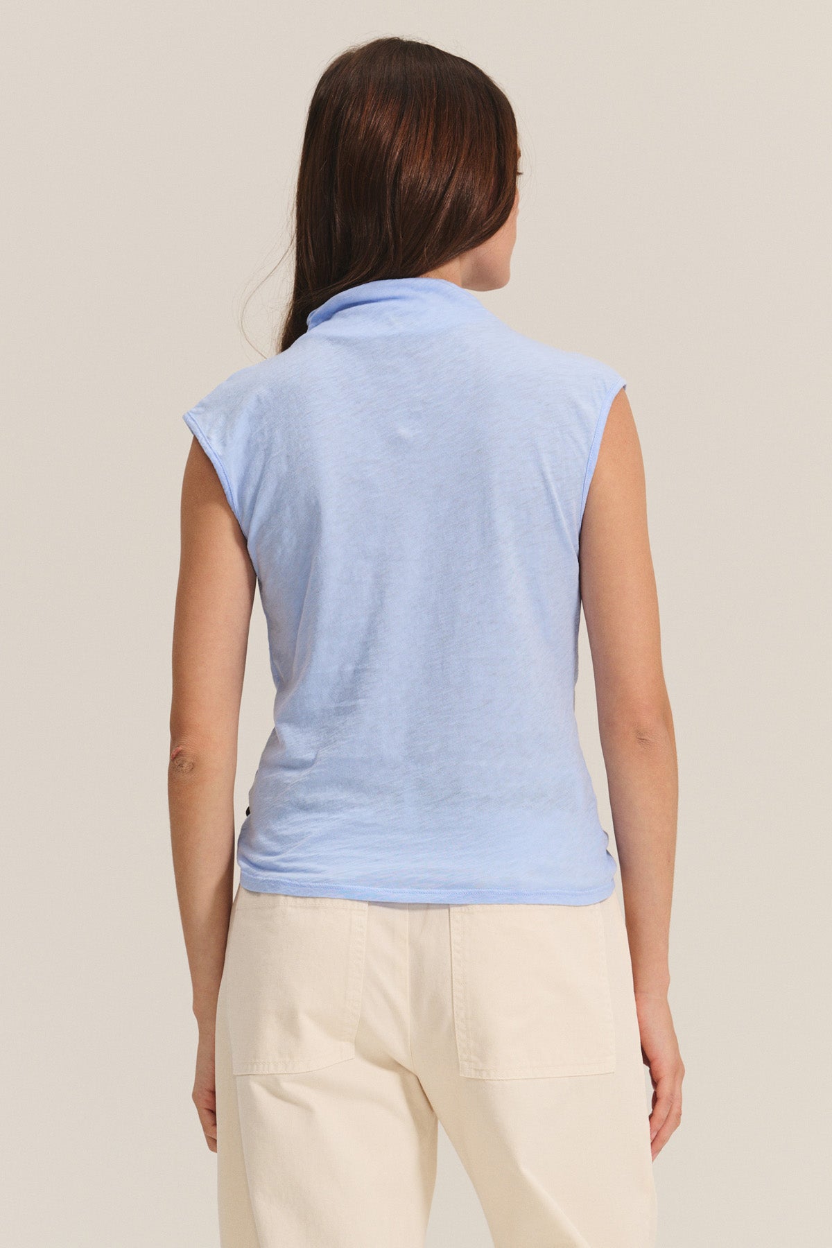 A woman with long brown hair is seen from the back against a plain background wearing the Velvet by Graham & Spencer FLETCHER TEE, a light blue mock neck sleeveless top, paired with cream-colored pants.-40681690497217