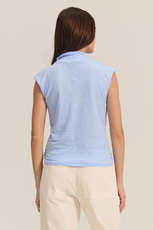 A woman with long brown hair is seen from the back against a plain background wearing the Velvet by Graham & Spencer FLETCHER TEE, a light blue mock neck sleeveless top, paired with cream-colored pants.
