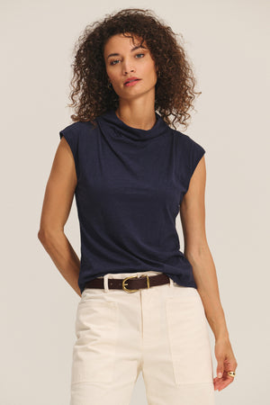 A woman with curly hair wears Velvet by Graham & Spencer’s FLETCHER TEE in navy, paired with high-waisted white pants and a brown belt, standing against a plain background.