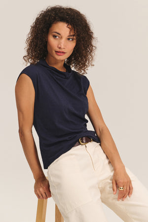 Someone with curly hair sits on a wooden stool against a neutral background, wearing the Velvet by Graham & Spencer FLETCHER TEE—a navy sleeveless top crafted from lightweight cotton slub—with light-colored pants.
