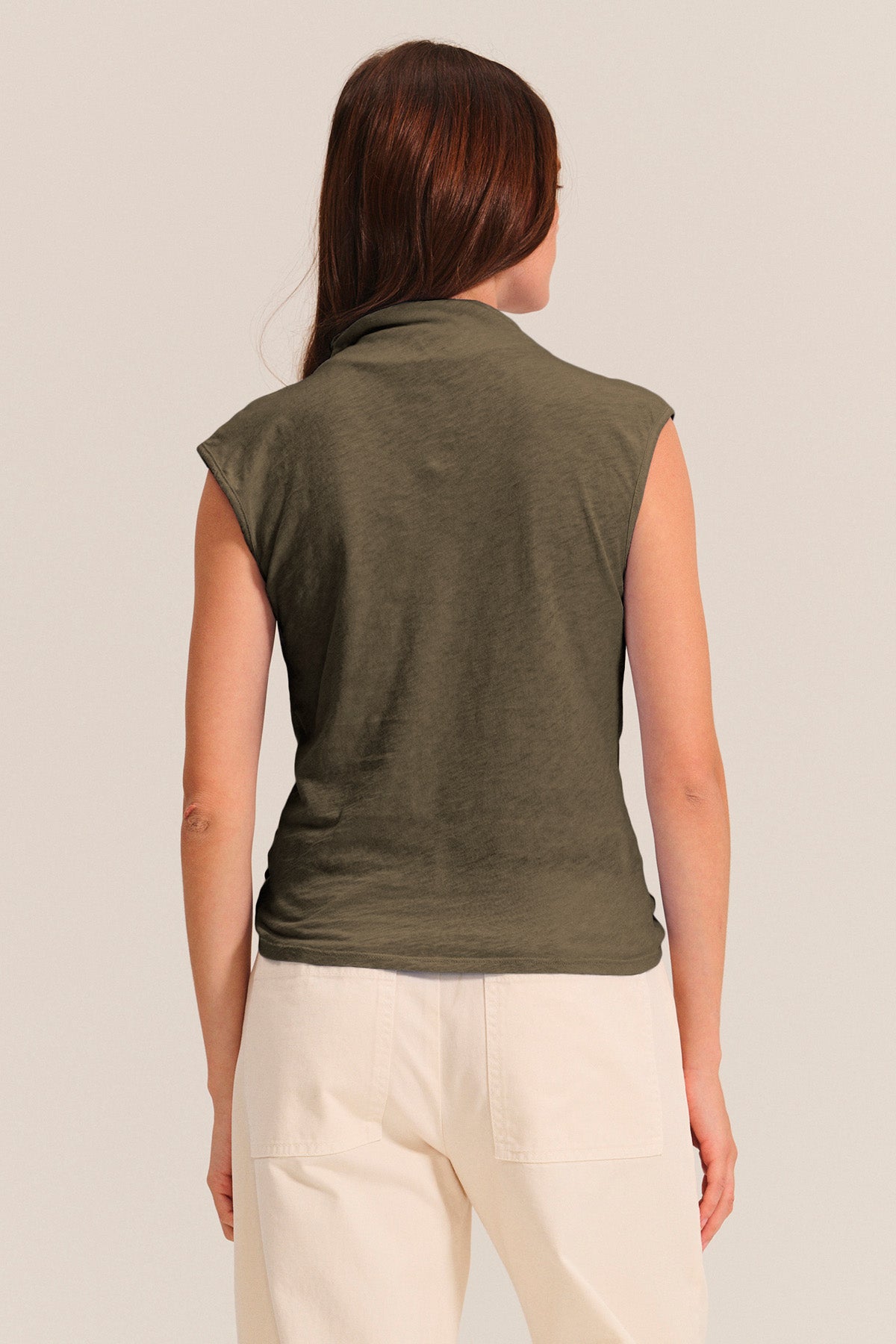 A woman with long brown hair is shown from the back, wearing the Velvet by Graham & Spencer FLETCHER TEE in olive green with white pants against a plain light background.