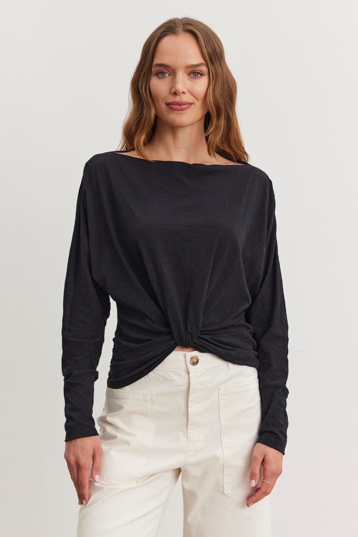 A woman with long hair wears the HALLIE TEE by Velvet by Graham & Spencer, a black top with a twist knot detail and relaxed dolman sleeves, paired with white pants, against a plain background.-38512533602497