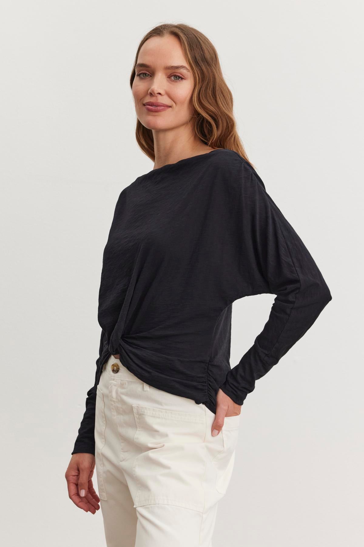   A person with long hair wears the HALLIE TEE by Velvet by Graham & Spencer, a black long-sleeve top with relaxed dolman sleeves, paired with white pants, standing and smiling against a plain background. 