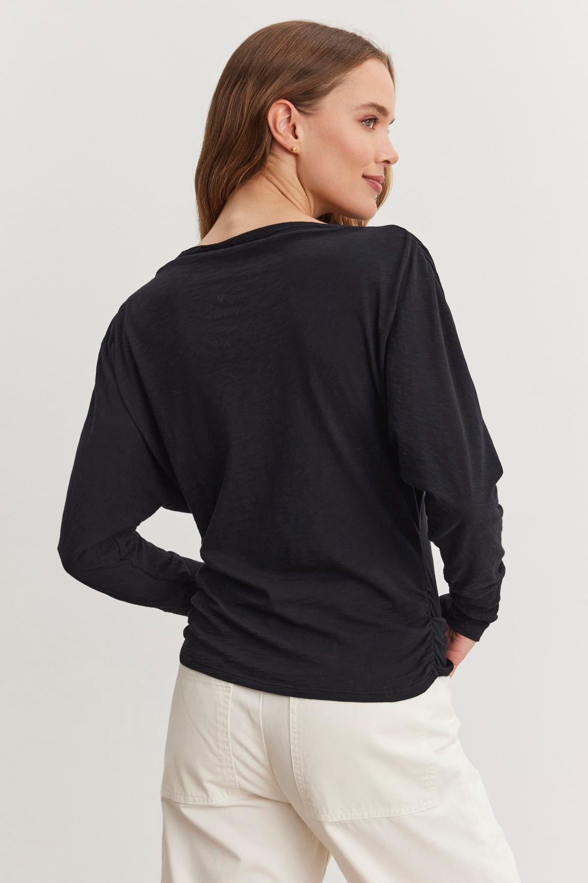 A person with long hair wears the HALLIE TEE by Velvet by Graham & Spencer, featuring relaxed dolman sleeves in black, and pairs it with white pants. They stand against a plain background, facing slightly to the right.-38512533668033