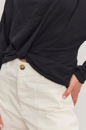 A person wearing the HALLIE TEE by Velvet by Graham & Spencer, featuring relaxed dolman sleeves and a twist knot detail, pairs it with white pants and casually places one hand in their pocket.