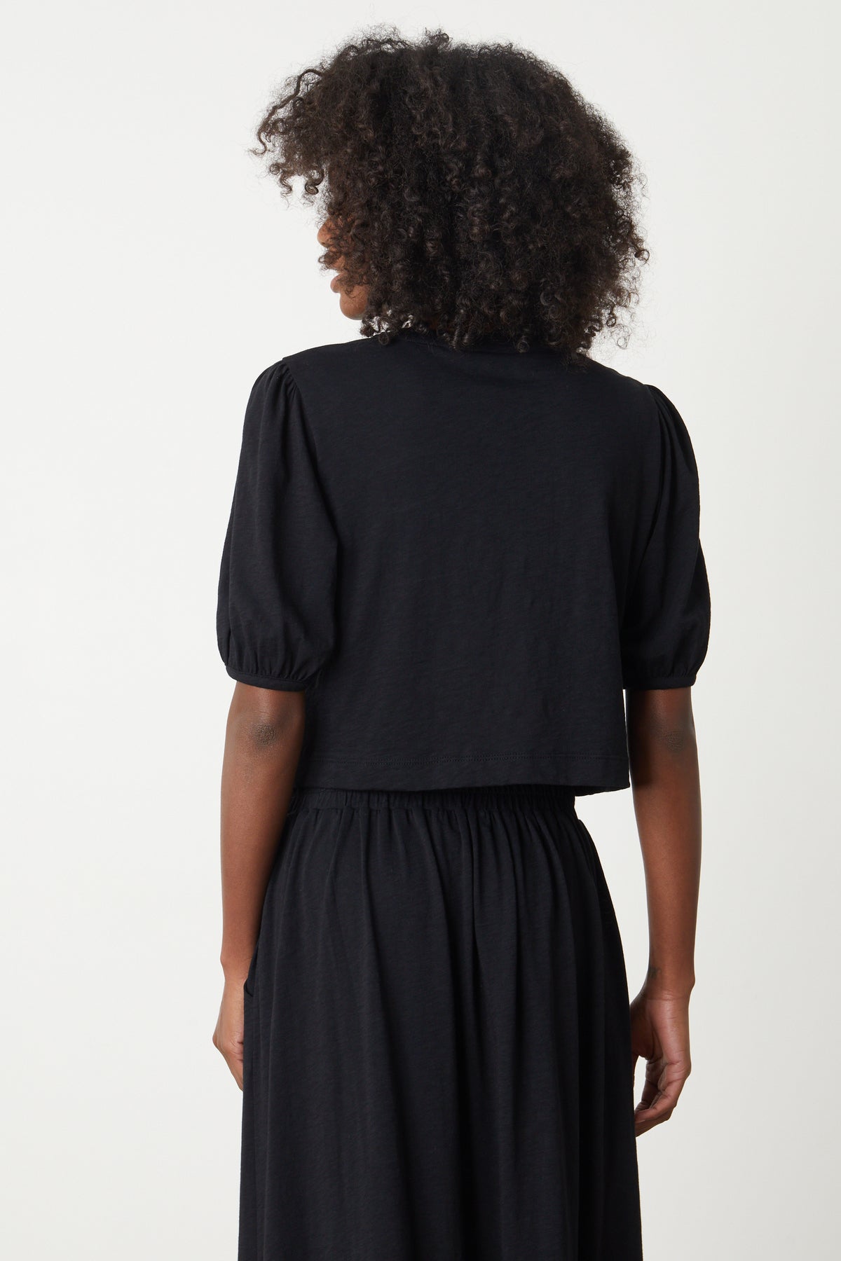   The back view of a woman wearing a black Velvet by Graham & Spencer Hilary Puff Sleeve Cropped Tee dress. 