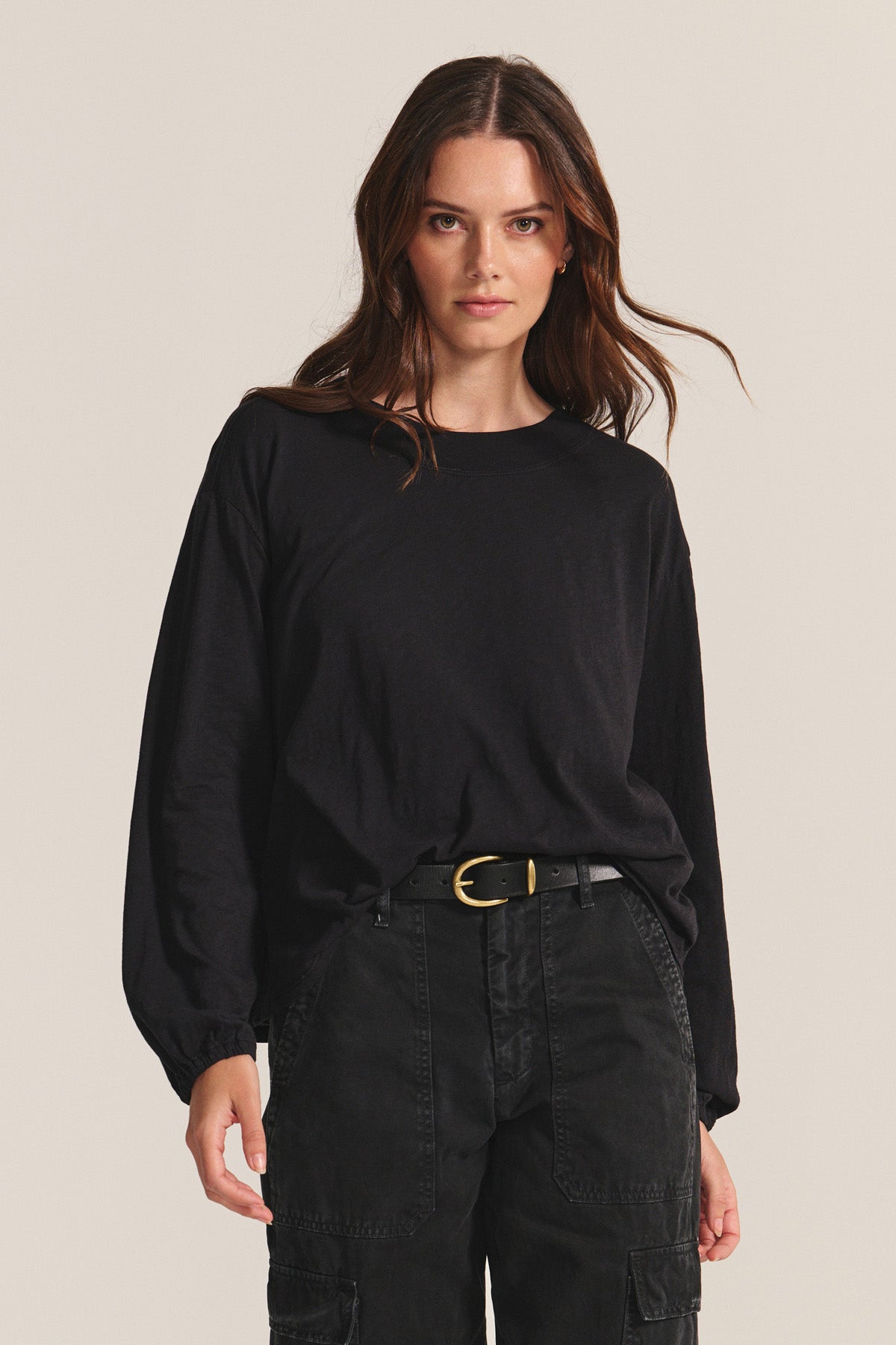 A woman with long brown hair wears the Velvet by Graham & Spencer JAMINA TEE in black, tucked into black cargo pants, standing against a plain light background.-40688692854977
