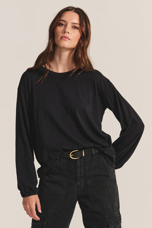 A woman wears the Velvet by Graham & Spencer JAMINA TEE, a loose-fitting black cotton slub top, tucked into black belted cargo pants against a beige background.