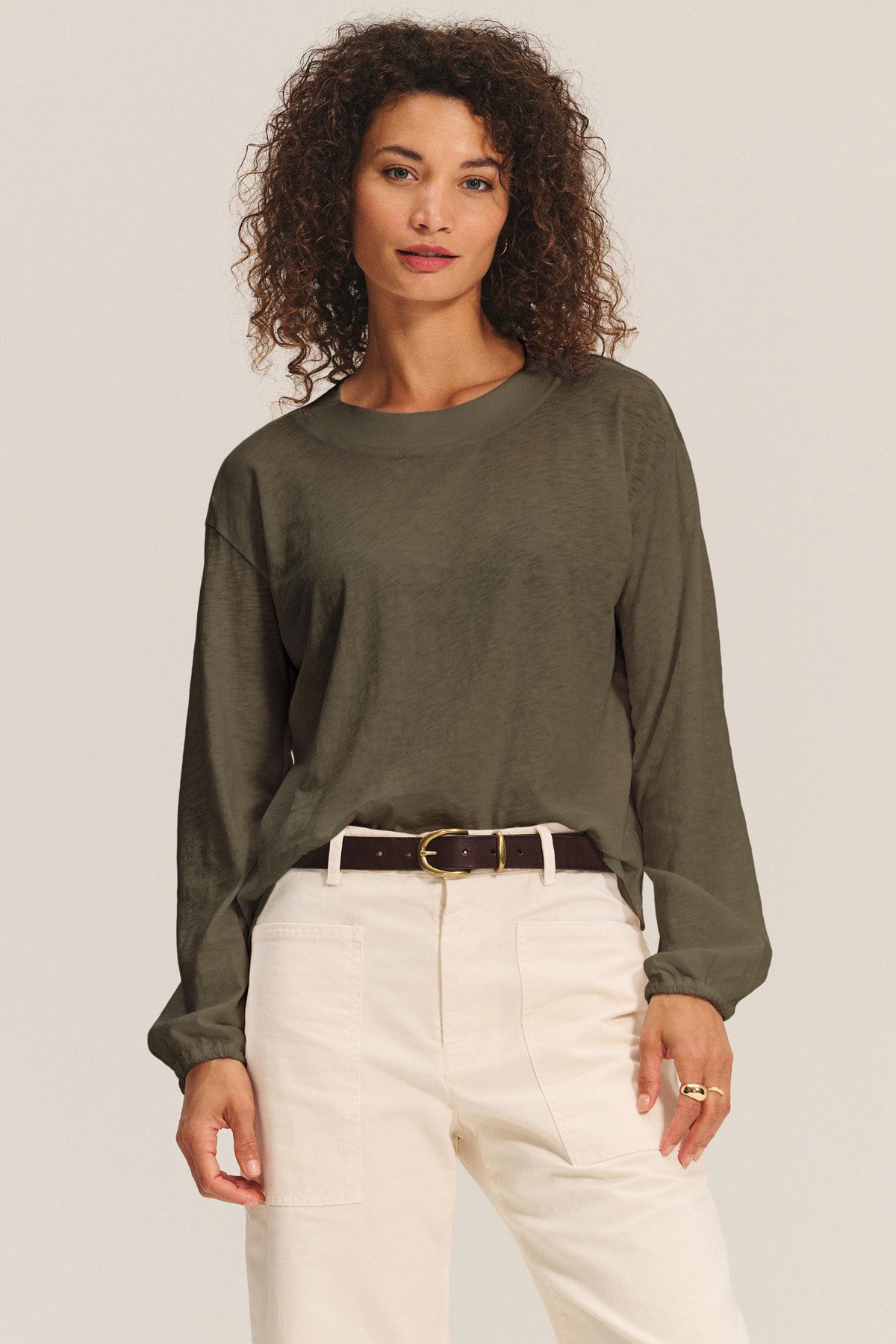 A woman stands against a neutral background wearing the JAMINA TEE by Velvet by Graham & Spencer—an olive cotton slub top tucked into high-waisted beige pants with a dark belt, creating an effortless everyday essential look.