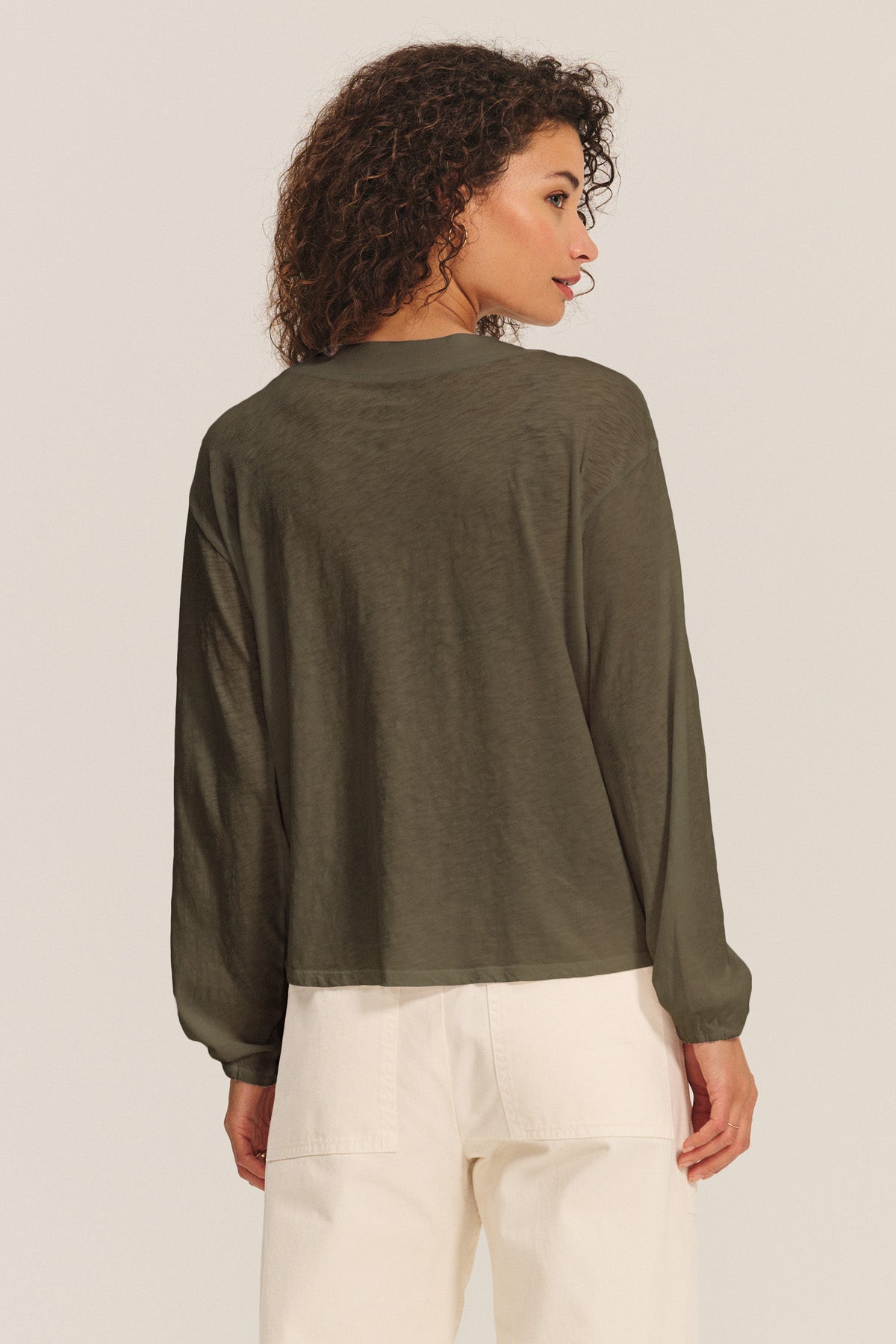 A person stands facing away in the Velvet by Graham & Spencer JAMINA TEE—a loose-fitting, long-sleeve cotton slub top—paired with high-waisted, off-white pants against a plain background.