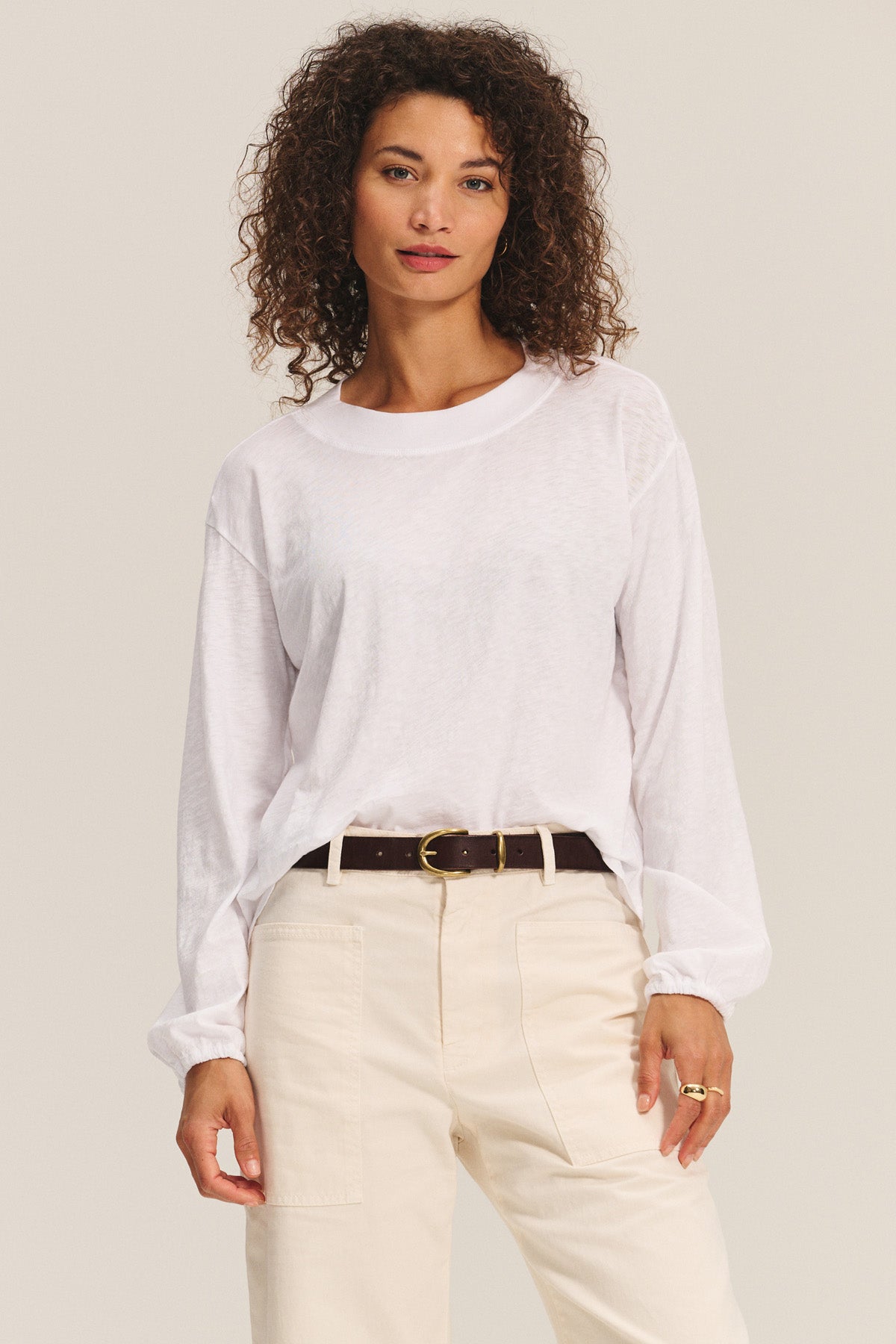 A woman with curly hair wears the JAMINA TEE by Velvet by Graham & Spencer—an essential white long-sleeve top—tucked into light beige pants with a black belt, standing against a plain background.-40732669608129