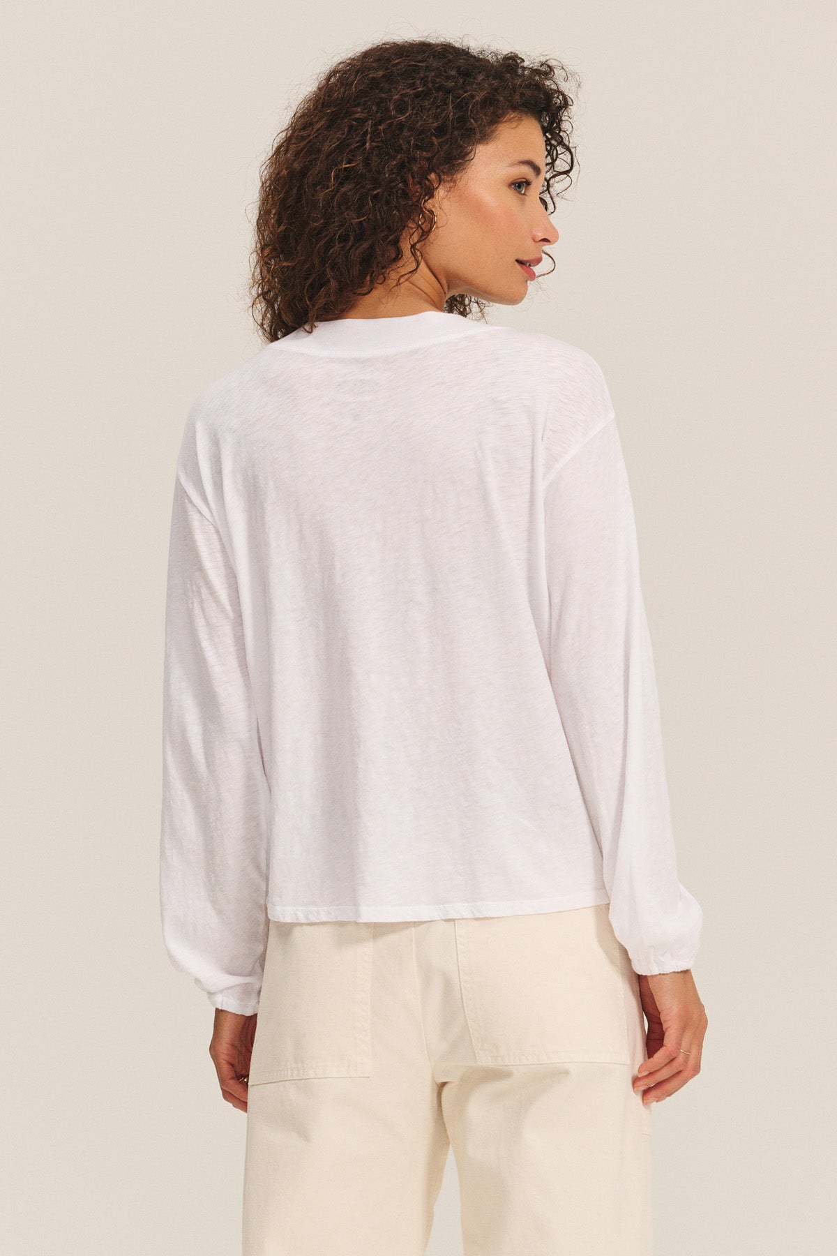 A woman stands with her back turned, wearing the JAMINA TEE by Velvet by Graham & Spencer—a long-sleeve cotton slub top paired with light pants—crafted as an everyday essential for any wardrobe against a plain backdrop.-40732669575361