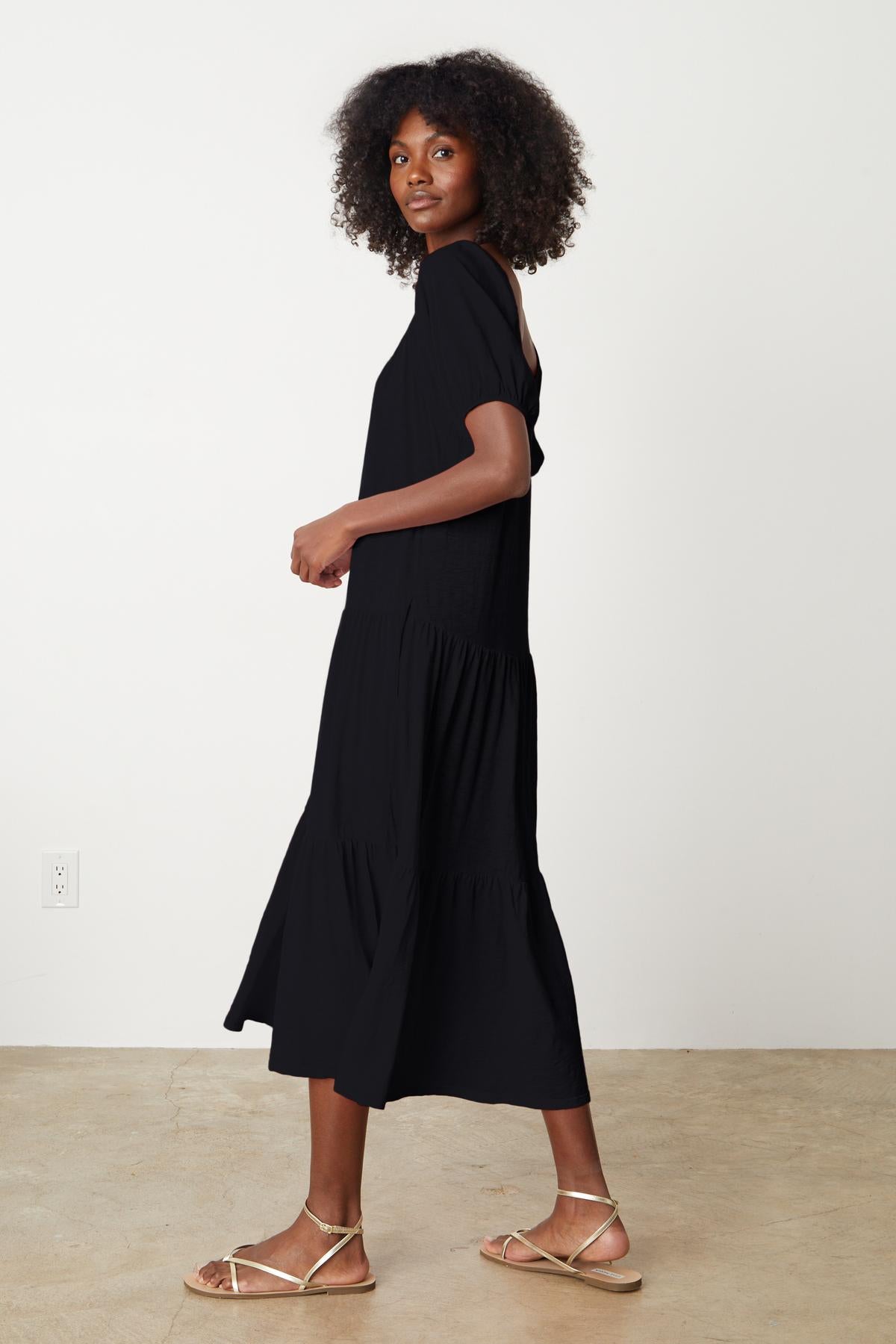   The model is wearing a black JANE SCOOP NECK TIERED DRESS by Velvet by Graham & Spencer and sandals. 
