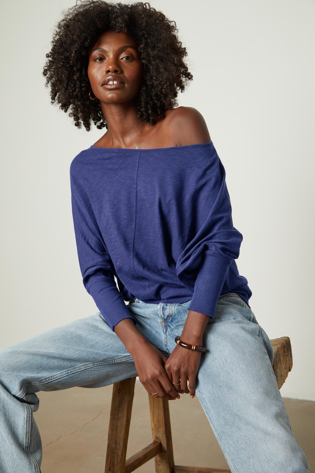   A woman is sitting on a stool wearing JOSS DOLMAN SLEEVE TEE by Velvet by Graham & Spencer jeans. 