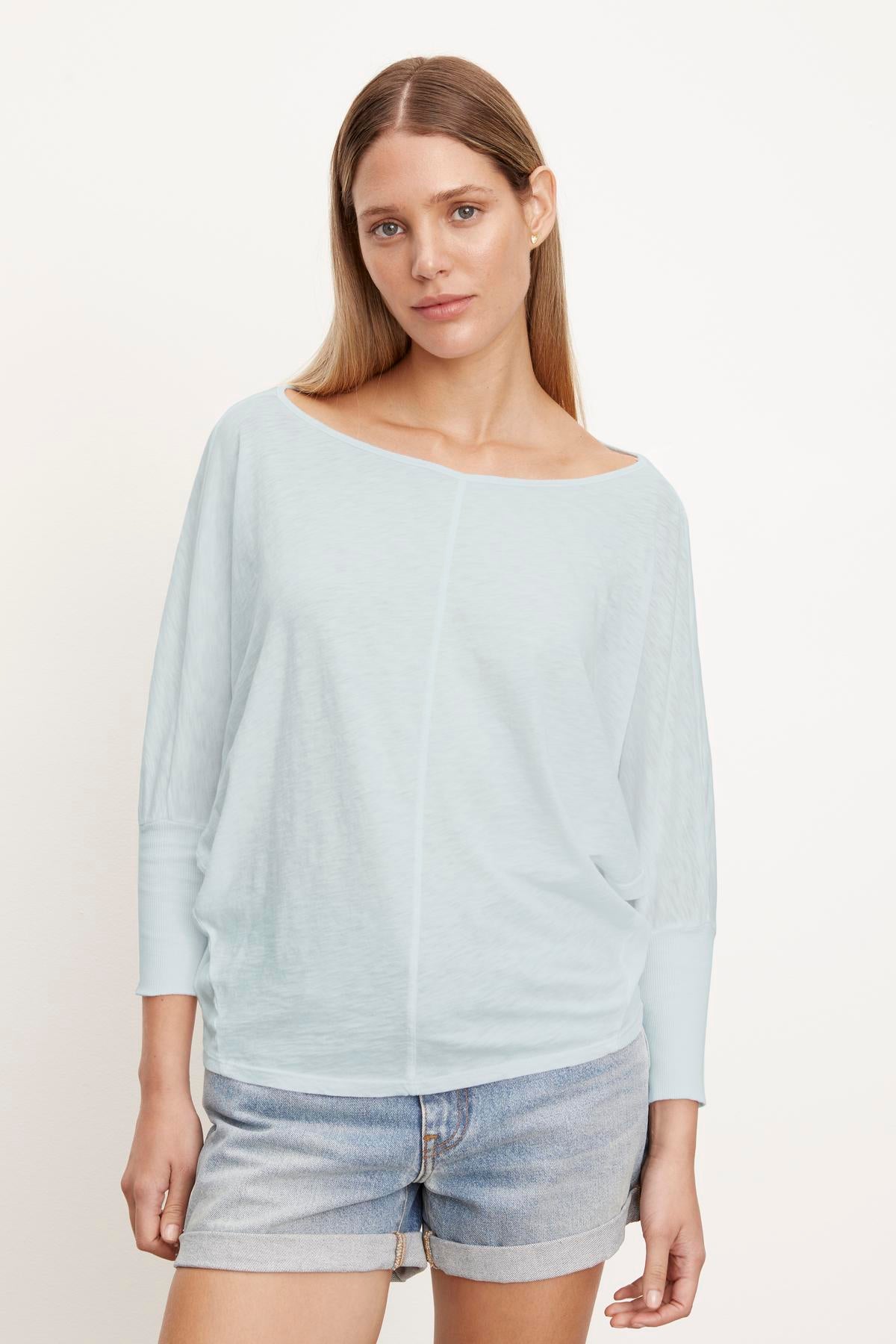 Woman with long brown hair wearing a light blue, slightly cropped JOSS DOLMAN SLEEVE TEE by Velvet by Graham & Spencer and denim shorts, standing in front of a plain white background.-37249861550273