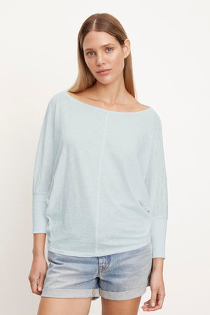 Woman with long brown hair wearing a light blue, slightly cropped JOSS DOLMAN SLEEVE TEE by Velvet by Graham & Spencer and denim shorts, standing in front of a plain white background.
