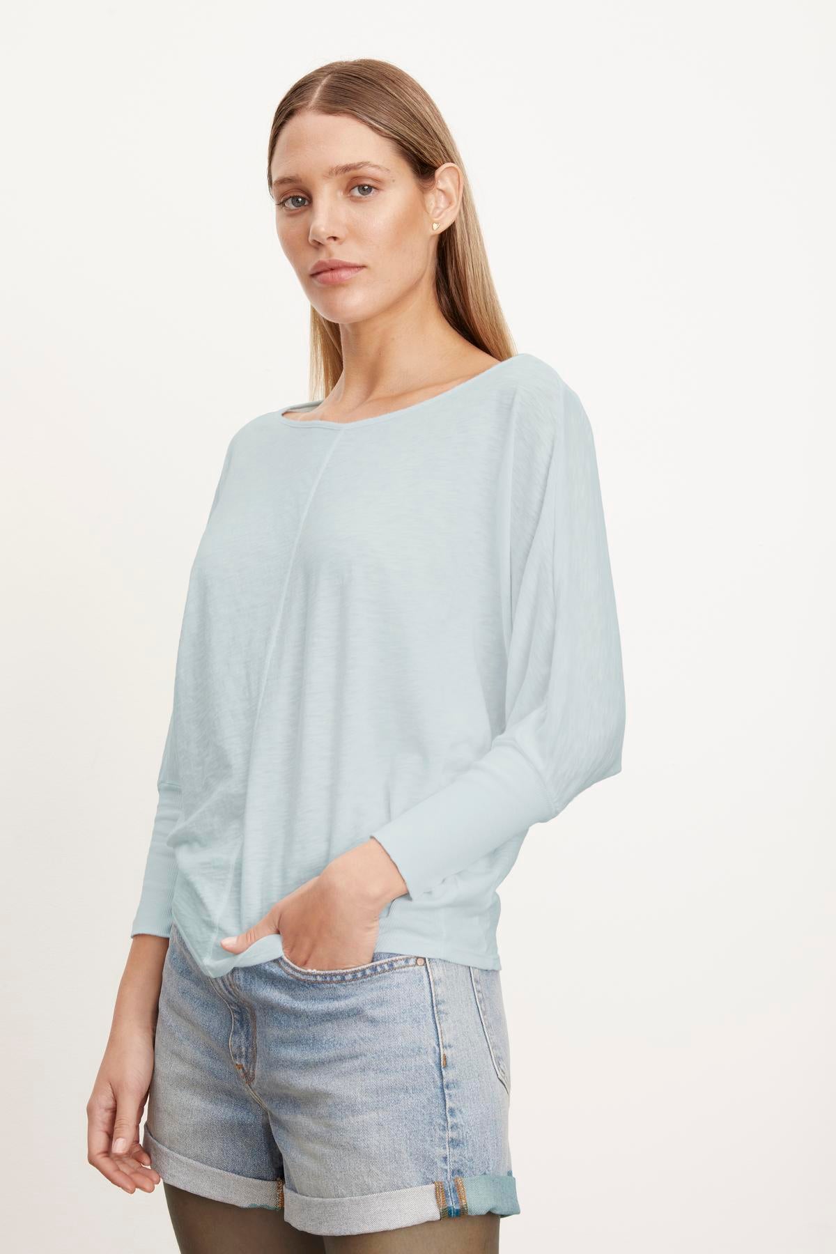 A person with long hair in a slightly cropped, light blue JOSS DOLMAN SLEEVE TEE by Velvet by Graham & Spencer and denim shorts stands against a plain background.-37249861517505