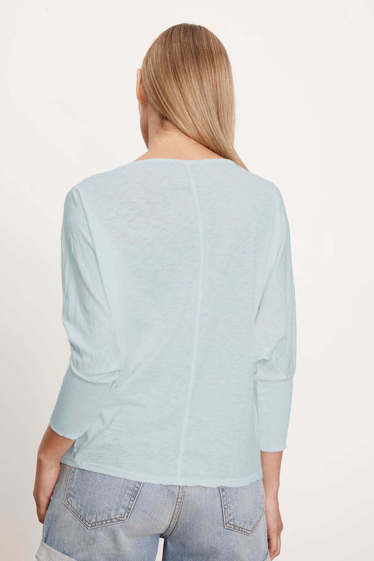 A woman with long blonde hair is shown from the back, wearing a light blue, slightly cropped Velvet by Graham & Spencer JOSS DOLMAN SLEEVE TEE and light-colored jeans.-37249861583041