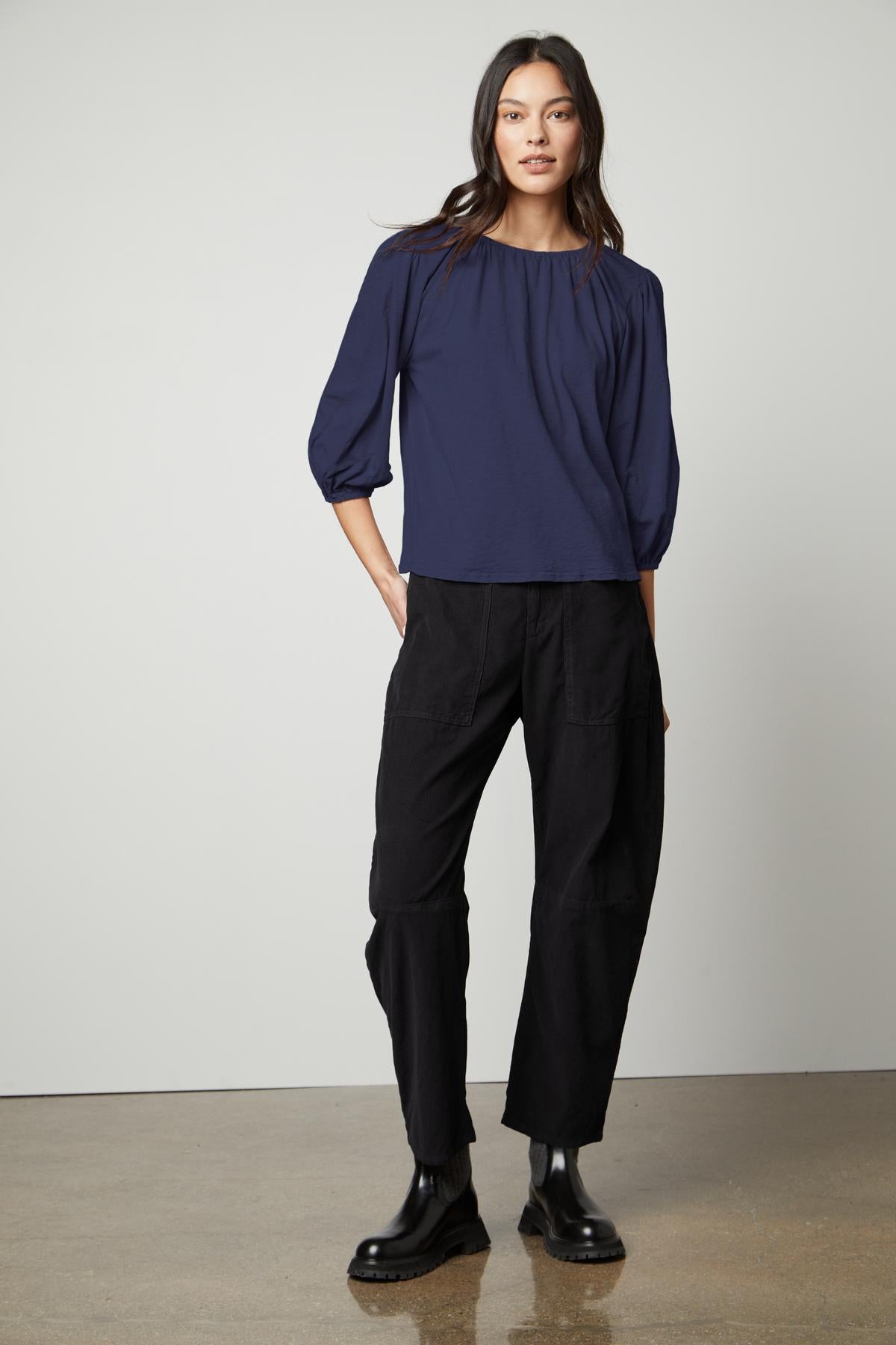   A woman stands against a plain background wearing the JULIE TEE by Velvet by Graham & Spencer, featuring puff sleeves and an elastic neckline in navy, paired with black pants and black boots. She has long dark hair and a neutral expression. 