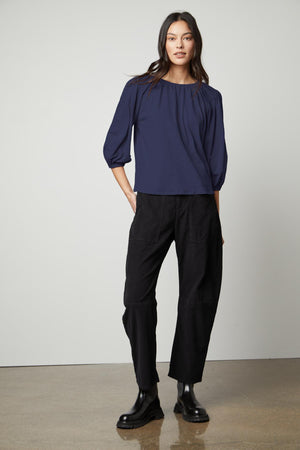 A woman stands against a plain background wearing the JULIE TEE by Velvet by Graham & Spencer, featuring puff sleeves and an elastic neckline in navy, paired with black pants and black boots. She has long dark hair and a neutral expression.