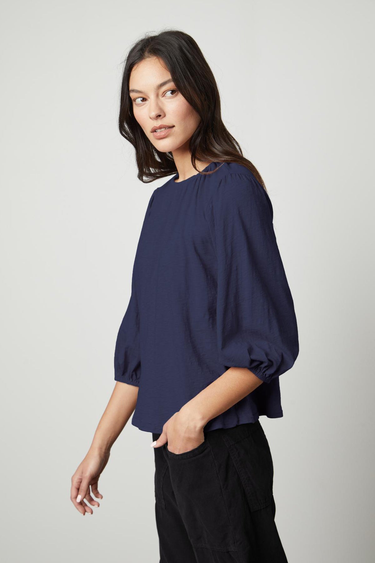   A woman with long dark hair is wearing the JULIE TEE by Velvet by Graham & Spencer, featuring navy blue three-quarter puff sleeves, paired with black pants. She stands against a plain light gray background, looking slightly to the side. 