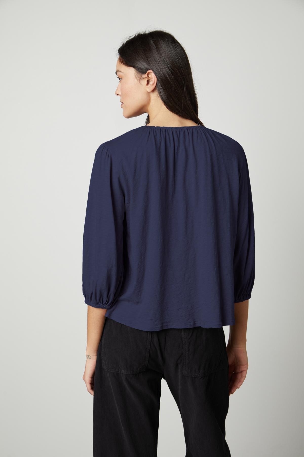   A person with long dark hair is shown from the back, wearing a navy blue JULIE TEE by Velvet by Graham & Spencer and black pants against a plain background. 