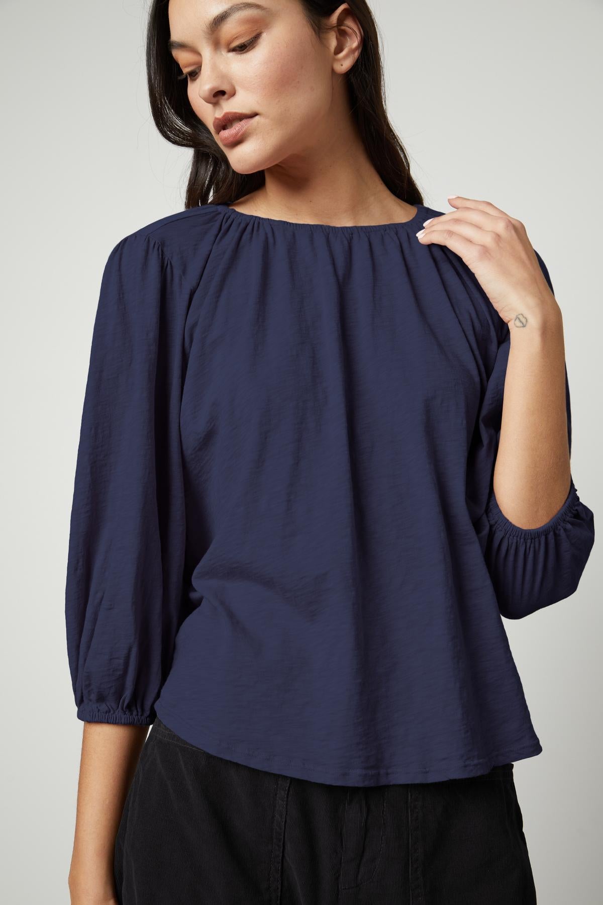   A person wearing the JULIE TEE, a blue long-sleeve blouse with puff sleeves and an elastic neckline by Velvet by Graham & Spencer, along with black pants, stands against a plain background. 