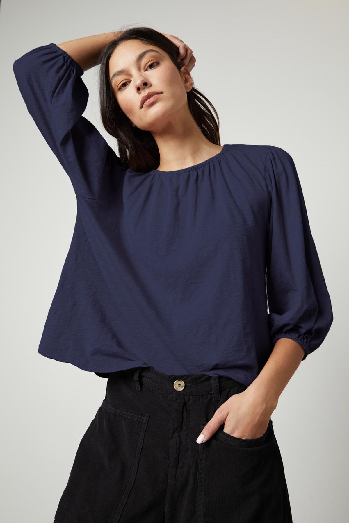   A person wearing the Velvet by Graham & Spencer JULIE TEE, a loose-fitting, long-sleeve dark blue blouse with an elastic neckline and puff sleeves, stands with one hand in their pocket and the other hand touching their head. 