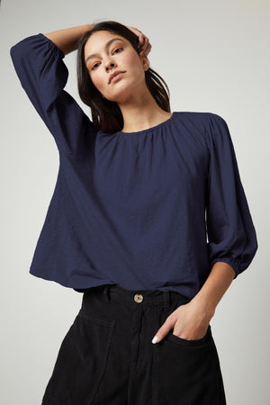 A person wearing the Velvet by Graham & Spencer JULIE TEE, a loose-fitting, long-sleeve dark blue blouse with an elastic neckline and puff sleeves, stands with one hand in their pocket and the other hand touching their head.