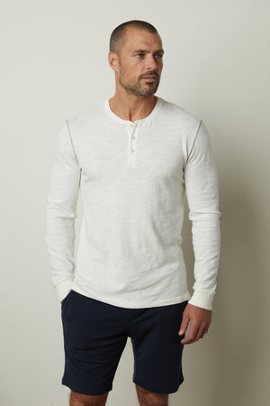 A man wearing a white Gabe Henley shirt by Velvet by Graham & Spencer in a cotton slub fabric, paired with blue shorts.