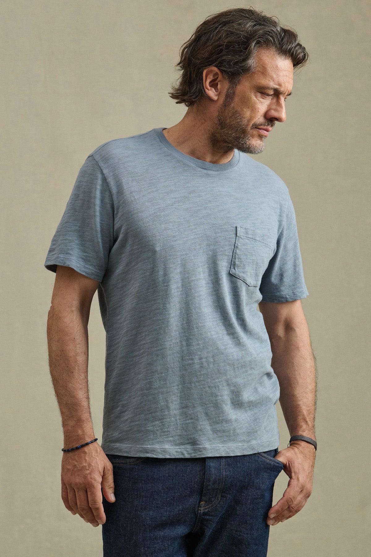 A man wearing the PAUL TEE by Velvet by Graham & Spencer, a light blue cotton slub crew neck with a chest pocket, paired with dark jeans, stands against a plain beige background.
