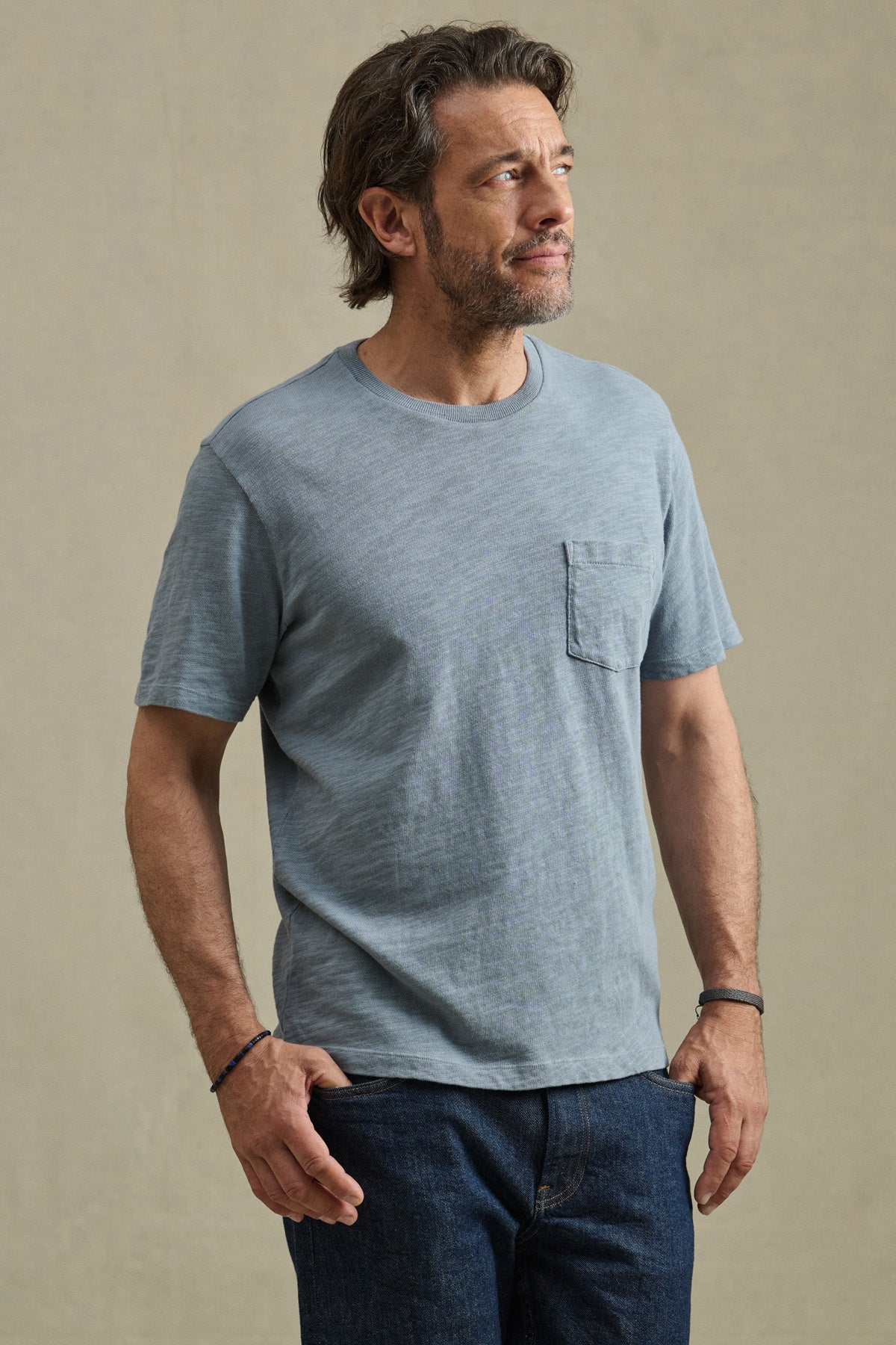 A man wears the PAUL TEE by Velvet by Graham & Spencer, a light blue crew neck short-sleeve cotton slub shirt with a chest pocket, paired with dark jeans, standing against a plain background and looking slightly aside.